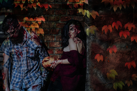 Halloween couple standing on brick wall with autumn leaves. Zombie man and woman with wounds and red blood. Girl holding bloody pumpkin. Halloween holiday celebration conceptの写真素材