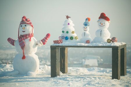 Snow sculptures on snowy landscape. Three snowmen in clothing on winter day outdoors. Christmas tree and present box on wooden table. xmas and new year. Holidays celebration conceptの写真素材