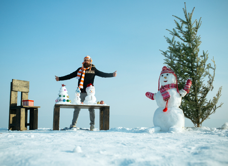 Man with snowy sculptures and presents on table and chair. Christmas and new year. Hipster enjoying winter day on blue sky. Holiday celebration concept. Snowman and xmas tree on snowy landscape.の写真素材