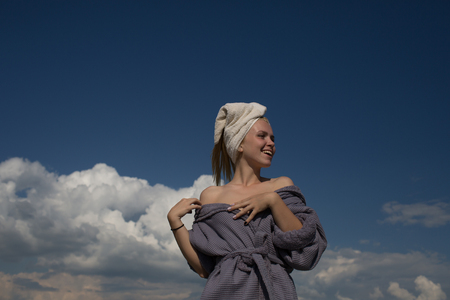 Girl smiling on cloudy blue sky. Spa and wellness. Happy model enjoying sunny day. Summer vacation concept. Woman in bathrobe and towel on head.の写真素材