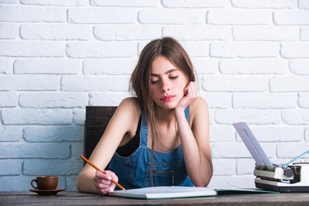 Study and learning concept. Education and knowledge. Girl writing with pencil in notebook. Young woman sitting at wooden table. Teenager student in jean pinafore on white brick wall.の写真素材