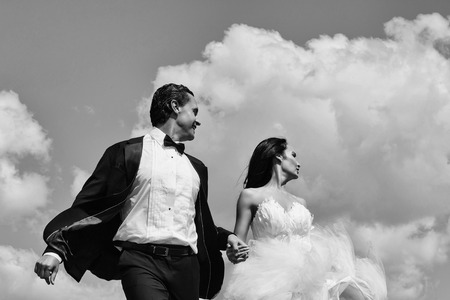 young wedding couple of sexy girl with brunette hair and pretty face in white bride dress and handsome man in black groom suit on cloudy blue sky natural backgroundの写真素材