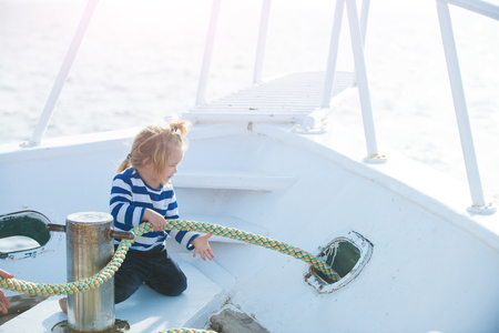 Travel and summer vacations. Baby care and childhood. Little child sitting and berthing rope on white boat. Boy kid playing with rope on yacht. Yachting and sailing concept.の写真素材
