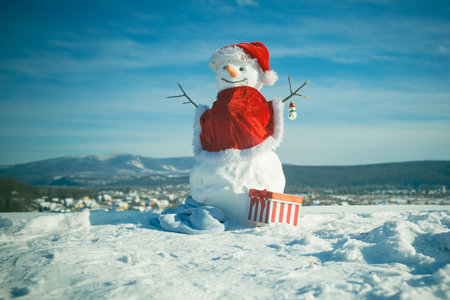 Snowman in red santa claus hat. Christmas or xmas decoration. New year snowman from white snow with gift box. Happy holiday and celebration. Winter activity and party.の写真素材