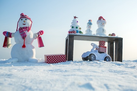 Snow sculptures and xmas tree on wooden table. Snowman with shopping bags on snowy background. Present boxes and toy car. Christmas and new year. Holidays celebration concept.の写真素材