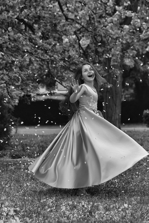 Beautiful little girl in glamour dress with long brunette hair and smiling happy face dancing with petals of spring pink flower blossom outdoorの写真素材