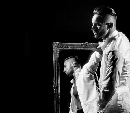 handsome muscular macho man or athlete guy with sexy body of bodybuilder with torso, belly, six packs, has beard on serious face in pants, white shirt near vintage mirror isolated on black backgroundの写真素材