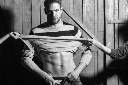 One sexual young stylish handsome muscular lover macho man with beard in sweater with bare torso and female mistress hand undressing him standing indoor in studio on wooden backdrop, horizontal photoの写真素材