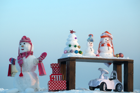 Snowman with shopping bags on blue sky. Snow sculptures and xmas tree on wooden table. Present boxes and toy car on snowy landscape. Christmas and new year. Holidays celebration concept.の写真素材