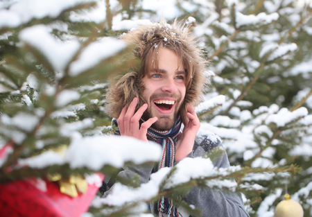 Guy smiling on fir trees on snowy nature. Macho with bearded face in snow forest. Christmas and new year. Holidays celebration concept. Happy man wearing fur hood and scarf on winter day.の写真素材