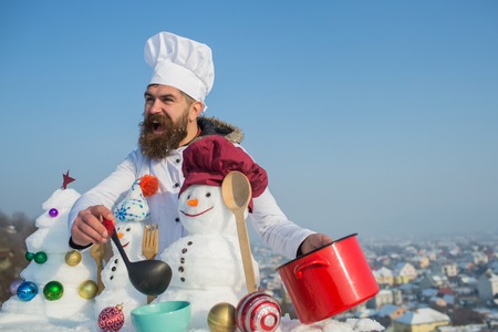 Excited man in chef hat ladling soup on winter day. Cook, snowmen and snow xmas tree on blue sky. Christmas and new year holidays food. Cooking and diet concept. Hipster with ladle in white uniform.の写真素材