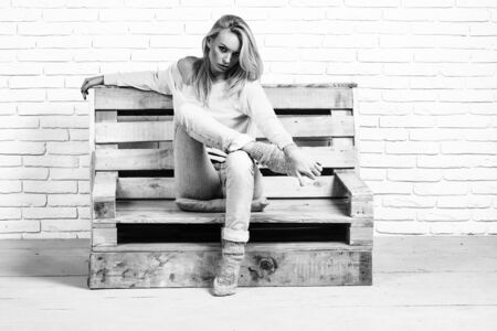girl or beautiful cute woman sexy fashion model with blond hair in jeans casual clothes sits on wooden pallet sofa on brick wall, black and whiteの写真素材