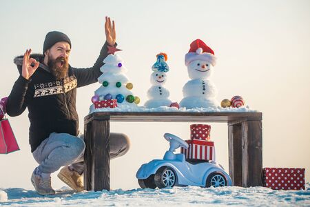 Man showing ok hand gesture. Happy hipster on white sky. Snowmen and xmas tree on wooden table. Toy car with gift boxes on snow. Christmas and new year presents concept.の写真素材