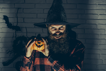 Halloween man with pumpkin in darkness. Scary hipster with beard in witch hat and plaid shirt. Holiday celebration symbols on brick wall. Mystery and horror concept. Jack o lantern.の写真素材