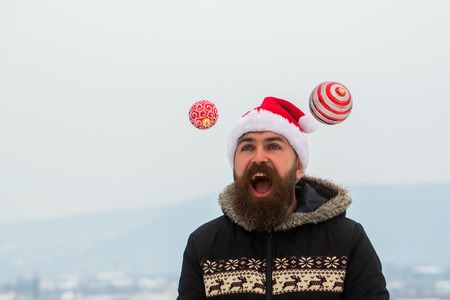 Happy hipster opening mouth with joy. Santa man juggling xmas balls on snowy landscape. Christmas and new year celebration. Guy in red santa claus hat and coat. Winter holidays concept.の写真素材