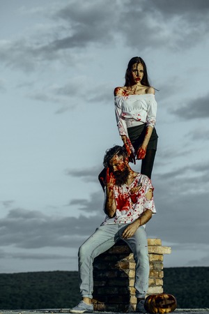 Halloween couple and pumpkin on chimney. Woman and man on grey sky. Vampires bleeding with red blood. Girl and hipster in bloody clothes on natural landscape. Halloween holiday conceptの写真素材