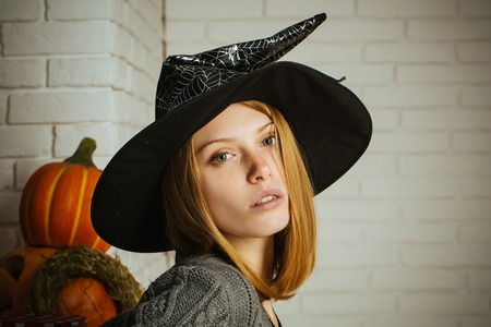 Halloween woman in witch hat. Girl with red hair and no makeup on face. Pumpkins and straw wreath on white brick wall. Holiday celebration concept. Tradition and traditional symbols.の写真素材