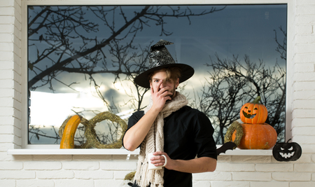 Halloween warm and homely atmosphere. Macho covering face in witch hat and scarf. Holiday symbols and decorations. Magic potion concept. Man holding cup at window with autumn trees.の写真素材