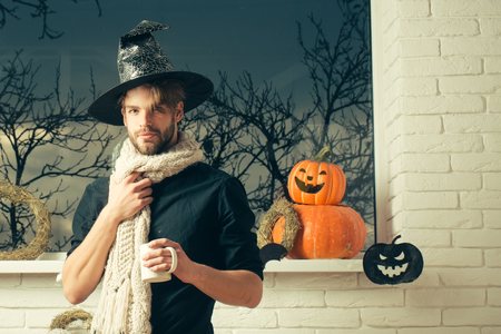Halloween holiday symbols and decorations. Man holding cup at window with autumn trees. Magic potion concept. Macho wearing witch hat and scarf. Warm and homely atmosphere.の写真素材