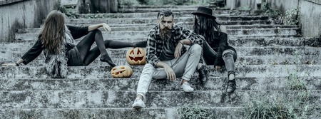 Halloween people and jack o lanterns. Halloween riends on stony stairs. Bearded man sitting with girls. Women with pumpkin heads. Holiday celebration concept.の写真素材