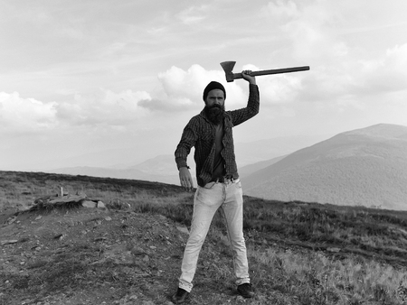 young handsome bearded hipster man in checkered shirt with long beard holding axe or ax on mountain top on natural background with skyの写真素材