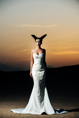 Woman posing on sunset sky. Girl in white wedding dress in desert. Bride in sand dunes on mountain landscape. Honeymoon travel concept. Summer vacation and holidays.の写真素材
