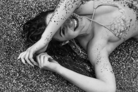 Pretty happy girl or woman, sexy brunette, dirty with natural sand lies in yellow swimsuit top on sea beach with transparent water outdoors on sunny summer day on grey sandy backgroundの写真素材