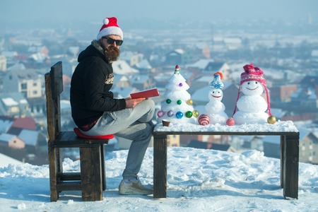 Hipster with book sitting on chair. Santa man reading on winter day. Christmas and new year holidays celebration. Snowmen and snow xmas tree on wooden table. Learning and fun concept.の写真素材