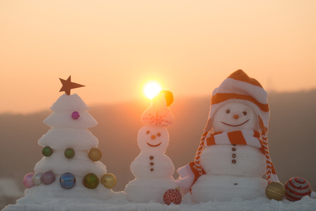 Snow sculptures on sunset sky background. xmas tree with star and balls. Winter holidays celebration concept. Merry Christmas and happy new year. Snowmen with smiley faces in hats on evening landscapeの写真素材