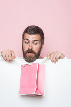 Guy with surprised face and gift pack. Hipster with present package on pink background. Christmas and birthday. Man with beard with pink shopping bag. Holiday celebration and black friday shopping.の写真素材