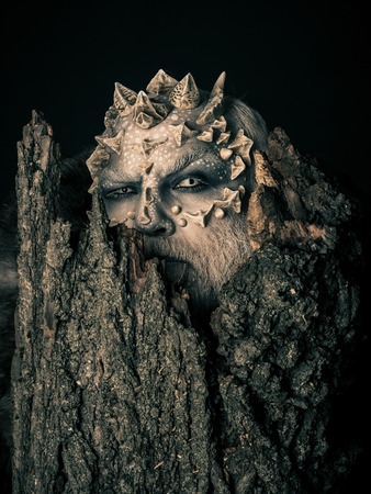Druid behind old bark on black background. Man with dragon skin and bearded face. Goblin with horns on head. Monster with sharp thorns and warts. Tree spirit and fantasy concept.の写真素材