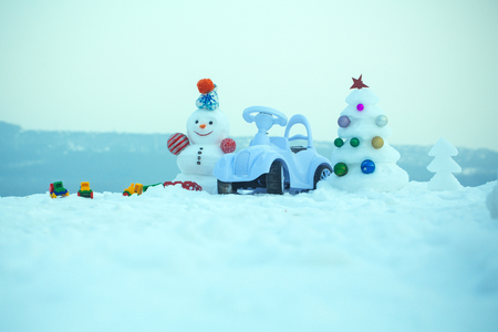 Snowman and toy car on snowy background. Holidays celebration concept. Christmas tree and present box on blue sky. Snow sculpture with smiley face on winter day. xmas and new year.の写真素材