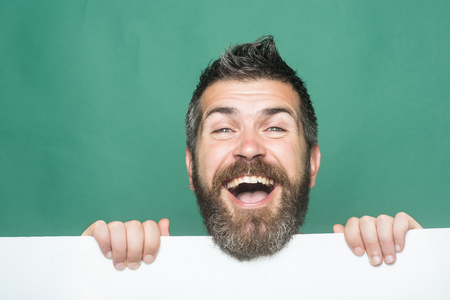 Guy or bearded man on green background. Feeling and emotions. Hipster with happy face hold paper. Man with long beard and mustache. Barber fashion and beauty marketing.の写真素材