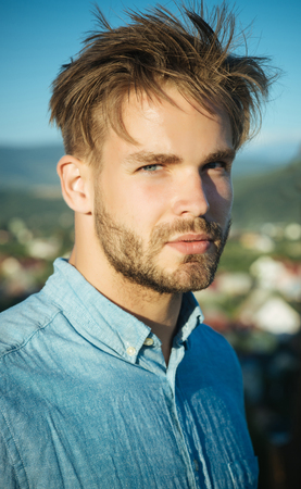 Man or macho with bearded face and stylish haircut posing in blue shirt on sunny day on natural landscape. Summer vacation and wanderlust. Male beauty concept.の写真素材