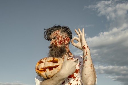 Halloween hipster holding bloody pumpkin on cloudy grey sky. Man bleeding with red blood and wounds. Vampire or zombie showing ok with jack o lantern. War, evil and horror. Holiday celebration conceptの写真素材