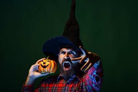 Halloween hipster with beard and jack o lantern. Man in wizard hat shouting with pumpkin on green background. Trick or treat. Autumn holiday celebration. Mystery and magic concept.の写真素材