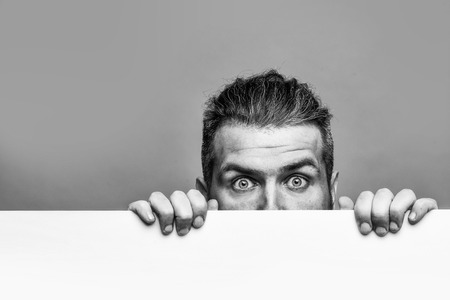 young man with scared eyes on emotional face with long hair behind white paper sheet in studio on grey background, copy spaceの写真素材