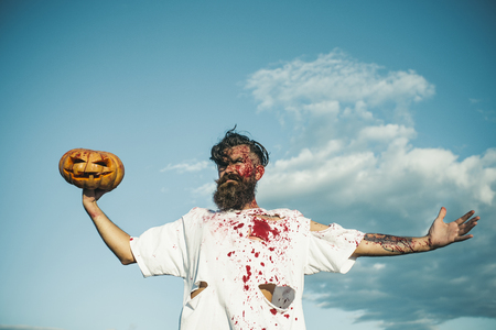 Halloween hipster in bloody torn tshirt with jack o lantern. War victim and horror concept. Autumn holiday celebration. Man holding pumpkin on hand. Soldier zombie with red blood on cloudy blue sky.の写真素材
