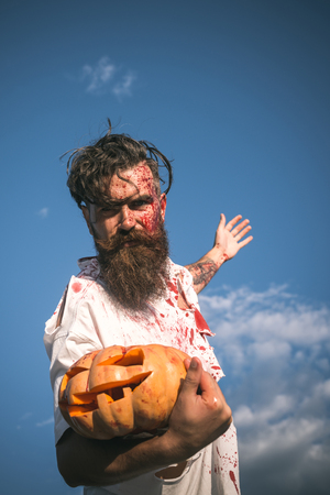 Halloween hipster in bloody torn tshirt with jack o lantern. Soldier zombie with red blood on cloudy blue sky. War victim and horror concept. Autumn holiday celebration. Man holding pumpkin on hand.の写真素材