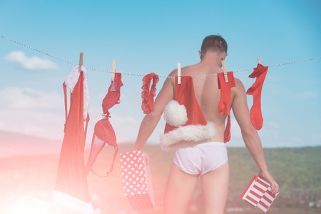 Santa claus man hanging clothes for drying. Laundry and dry cleaning. Xmas red costume on rope with pin. Christmas man hold present box. New year guy with muscular body on blue sky.の写真素材