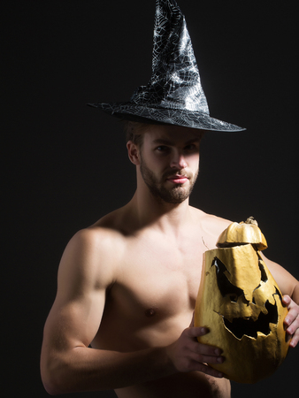 Halloween man in witch hat on black background. Macho with muscular torso, biceps, triceps holding pumpkin. Spirit and supernatural concept. Evil spell and magic. Autumn holidays and harvest seasonの写真素材