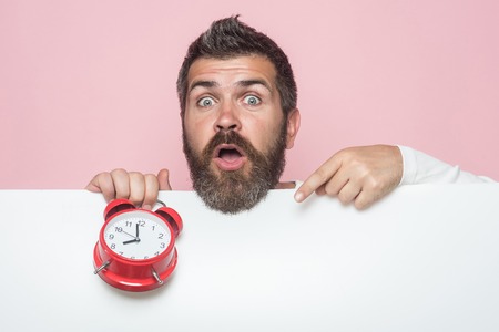 Man with long beard hold alarm clock. Hipster with red clock on pink background. End of working day or perfect morning. Guy with surprised face hold white paper. Time management and business success.の写真素材