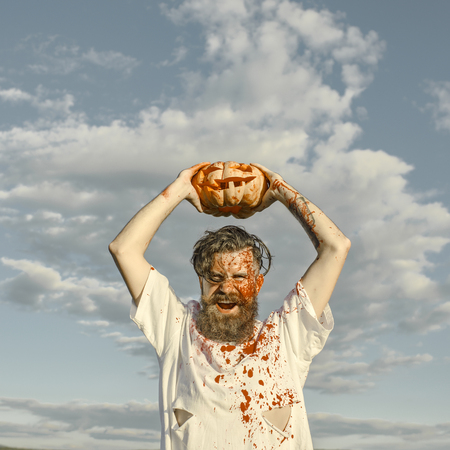 Halloween hipster with red blood shouting angry. Man holding pumpkin overhead on cloudy grey sky. Aggression, war concept. Soldier in bloody torn tshirt with jack o lantern. Autumn holiday celebrationの写真素材