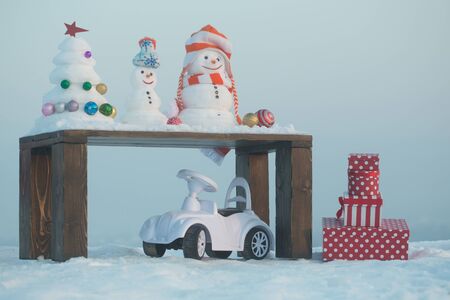 Winter holidays concept. Toy car with present boxes on snowy background. Snow sculptures on wooden table on blue sky. Christmas and new year celebration. Snowmen and xmas tree with baubles.の写真素材