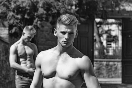 Young man handsome looks at camera while twin brother muscular bodybuilder with bare chest poses outdoor on blurred backgroundの写真素材