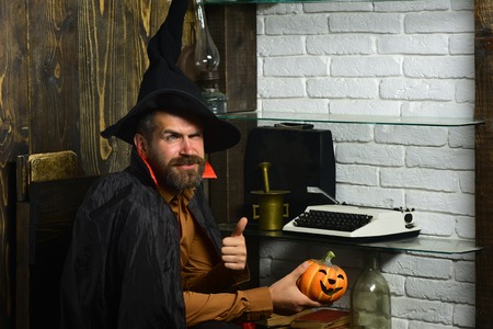 Halloween holiday celebration concept. Man in witch hat and cloak showing thumbs up. Wizardry and witchcraft. Wizard sitting with pumpkin on wooden wall. Magic and evil spell.の写真素材