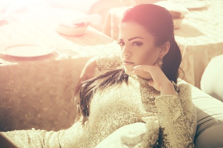 Woman in party dress with golden sequins relaxing on sofa in restaurant on sunny day. Holiday celebration and vacation conceptの写真素材