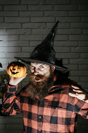 Halloween man with pumpkin in darkness. Holiday celebration symbols on brick wall. Jack o lantern. Mystery and horror concept. Hipster with frown face in witch hat and plaid shirt.の写真素材