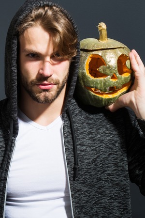 young handsome bearded sexy man with stylish hair in white shirt and jacket in studio on grey wall background holds orange golden halloween traditional pumpkin with scary face, copy spaceの写真素材