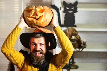 Wizard in black witch hat on spooky room background, defocused. Man with beard and happy face holds jack o lantern. Magician and carved pumpkin. Halloween decoration conceptの写真素材